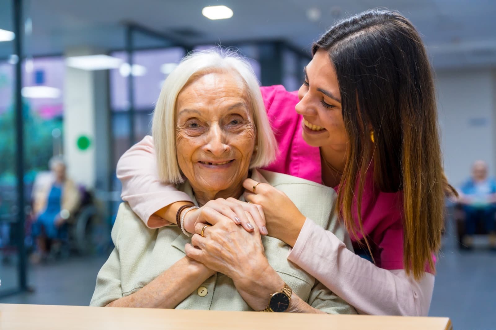 Cuidadora de uniforme rosa abrazando por detrás a una señora mayor sonriente